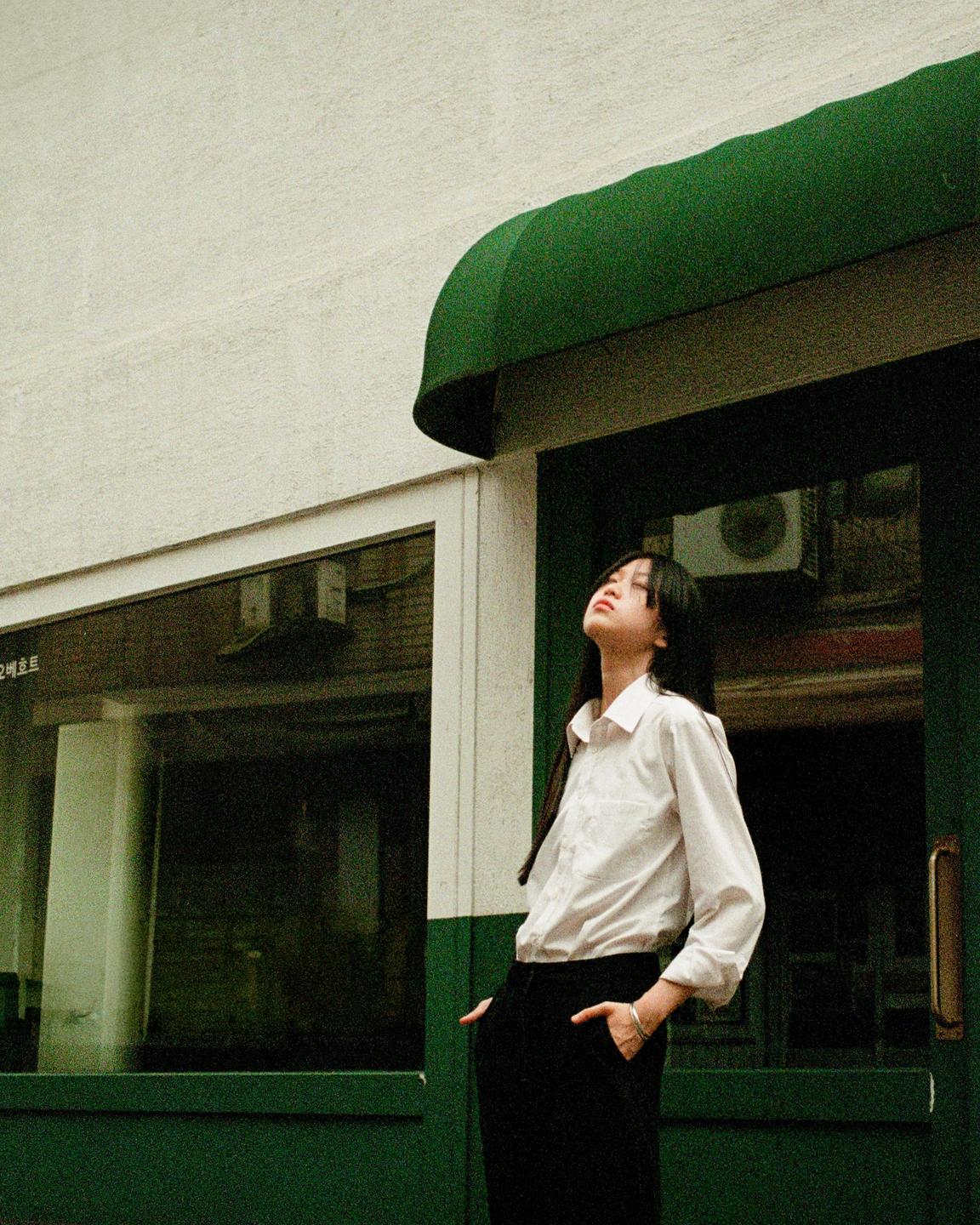 A woman stands with her head tilted back, looking up, next to a building with a green awning and trim.