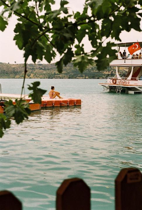 A woman sits on a floating dock next to a boat with a Turkish flag on a lake with hills in the background.
