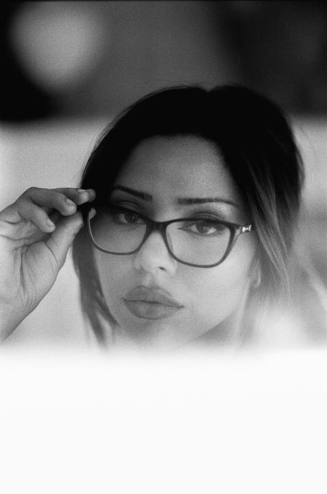 A woman with dark hair and glasses looks directly at the viewer, holding the frame of her glasses with one hand.