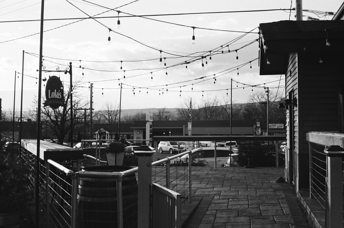A black and white street photograph shows a cafe sign and string lights hanging over a paved patio with cars and buildings in the background.