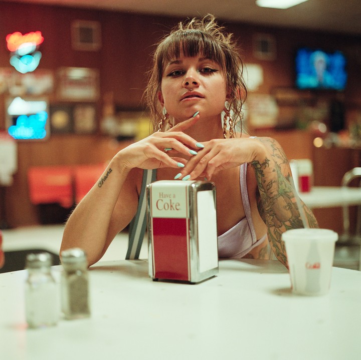 A woman with tattoos on her arms sits at a diner table with a napkin dispenser and salt and pepper shakers.