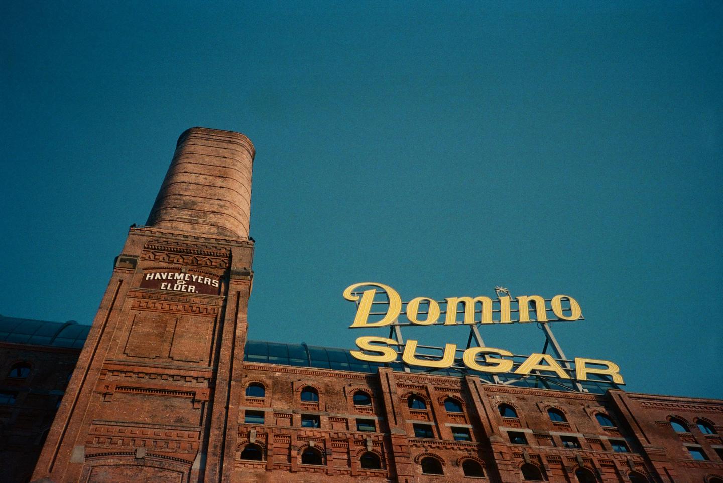 A brick building with a tall chimney and a large yellow sign that reads "Domino Sugar" is shown against a clear blue sky.