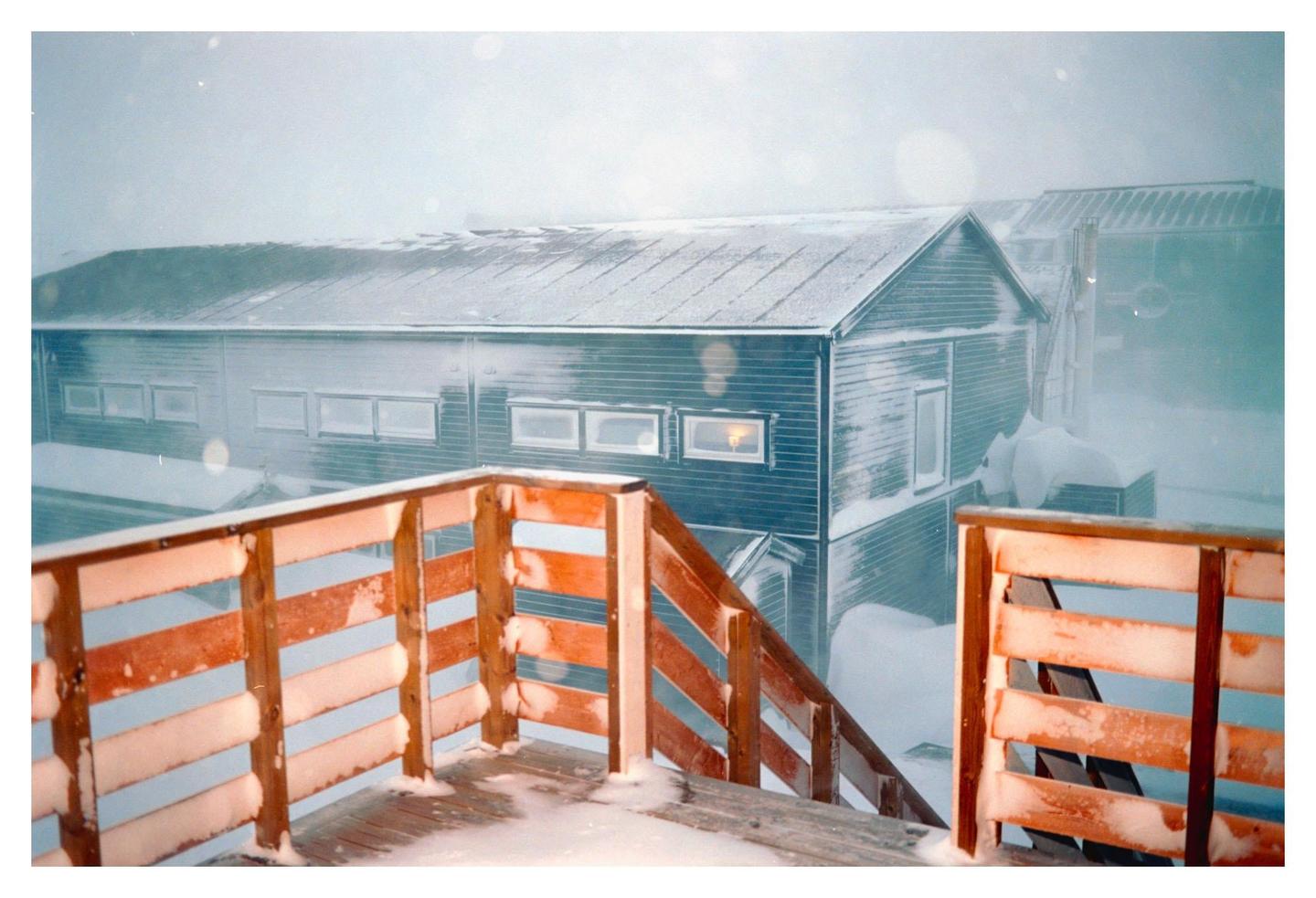 A snow-covered wooden deck and railing are in the foreground, with a building and snowdrifts in the background during a snowstorm.