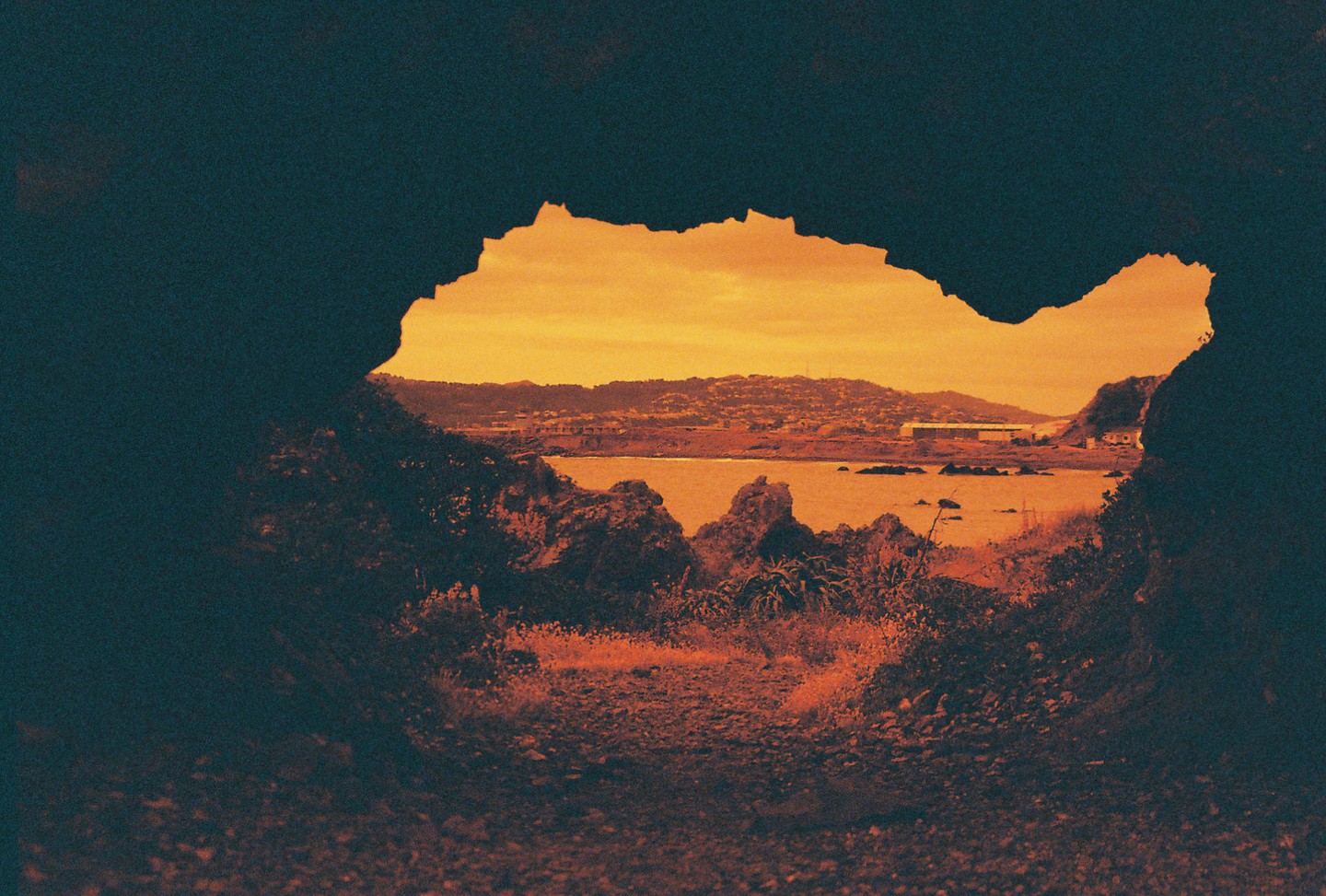 A view of a bay with hills and buildings in the distance is framed by a dark, jagged opening in the foreground.