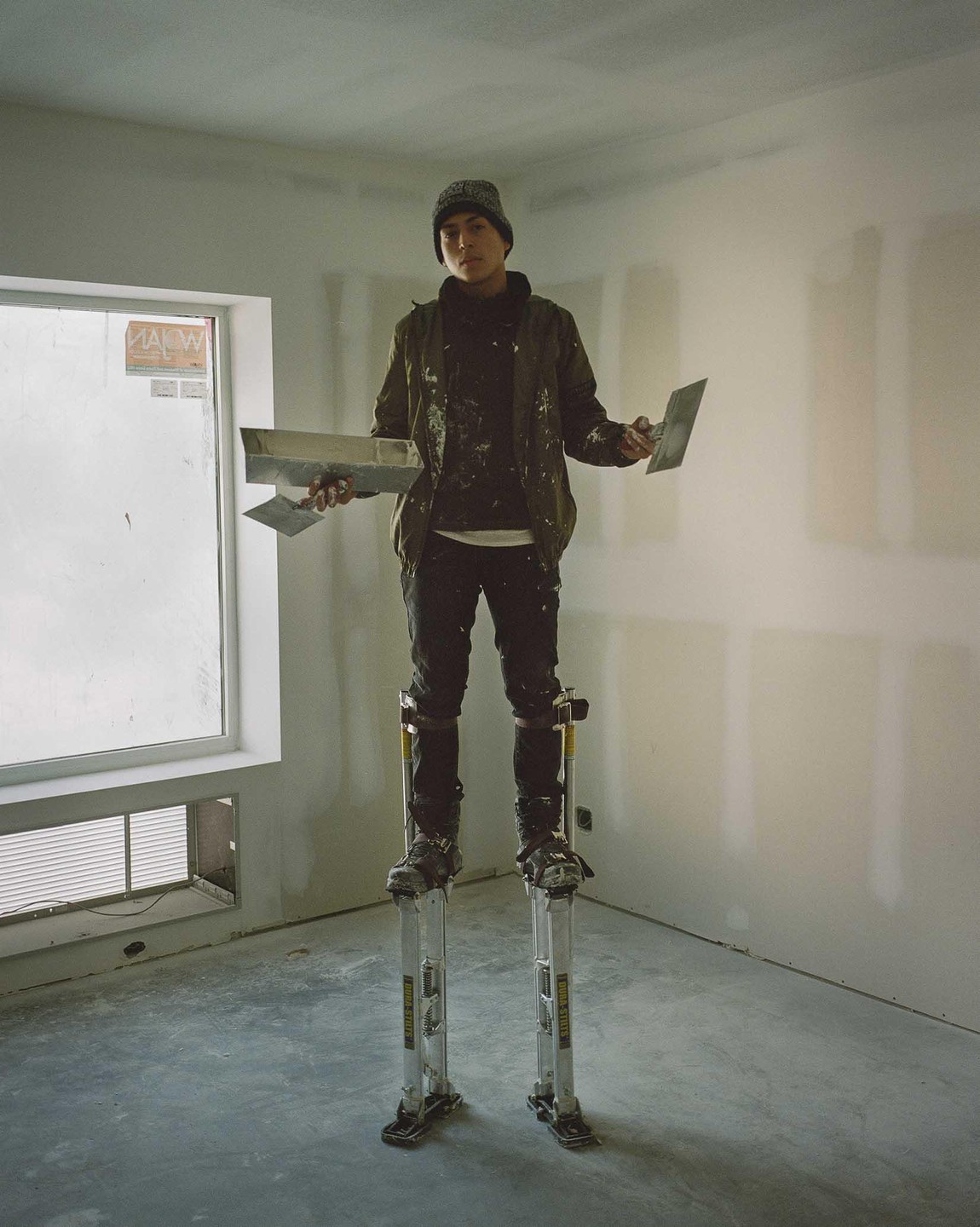 A man wearing stilts and holding drywall tools stands in a room with unfinished walls.