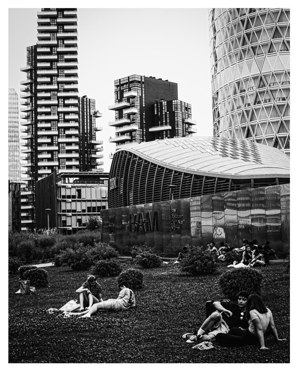 People relax on a grassy area surrounded by modern buildings in Milan.