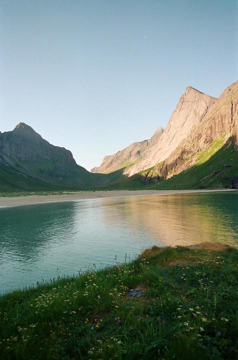 A body of water reflects the sky and mountains, with a sandy beach and grassy bank in the foreground.