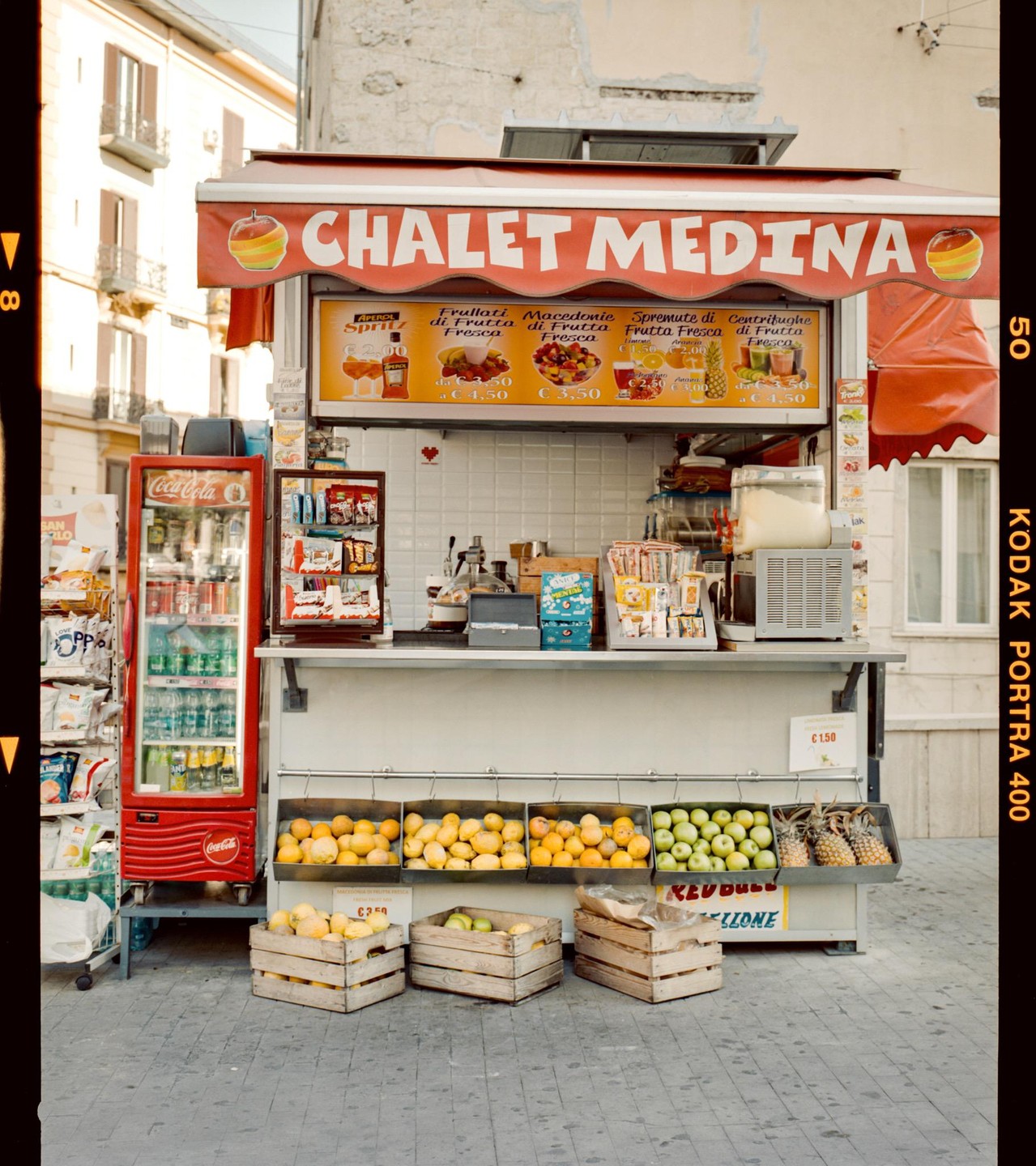 A fruit stand with a red awning displays various fruits and drinks on a city street.