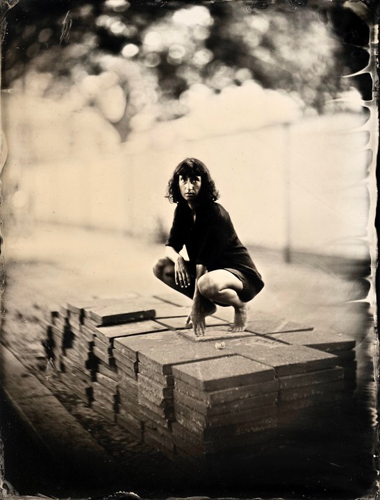 A woman crouches barefoot on a stack of square tiles in a sepia-toned portrait.