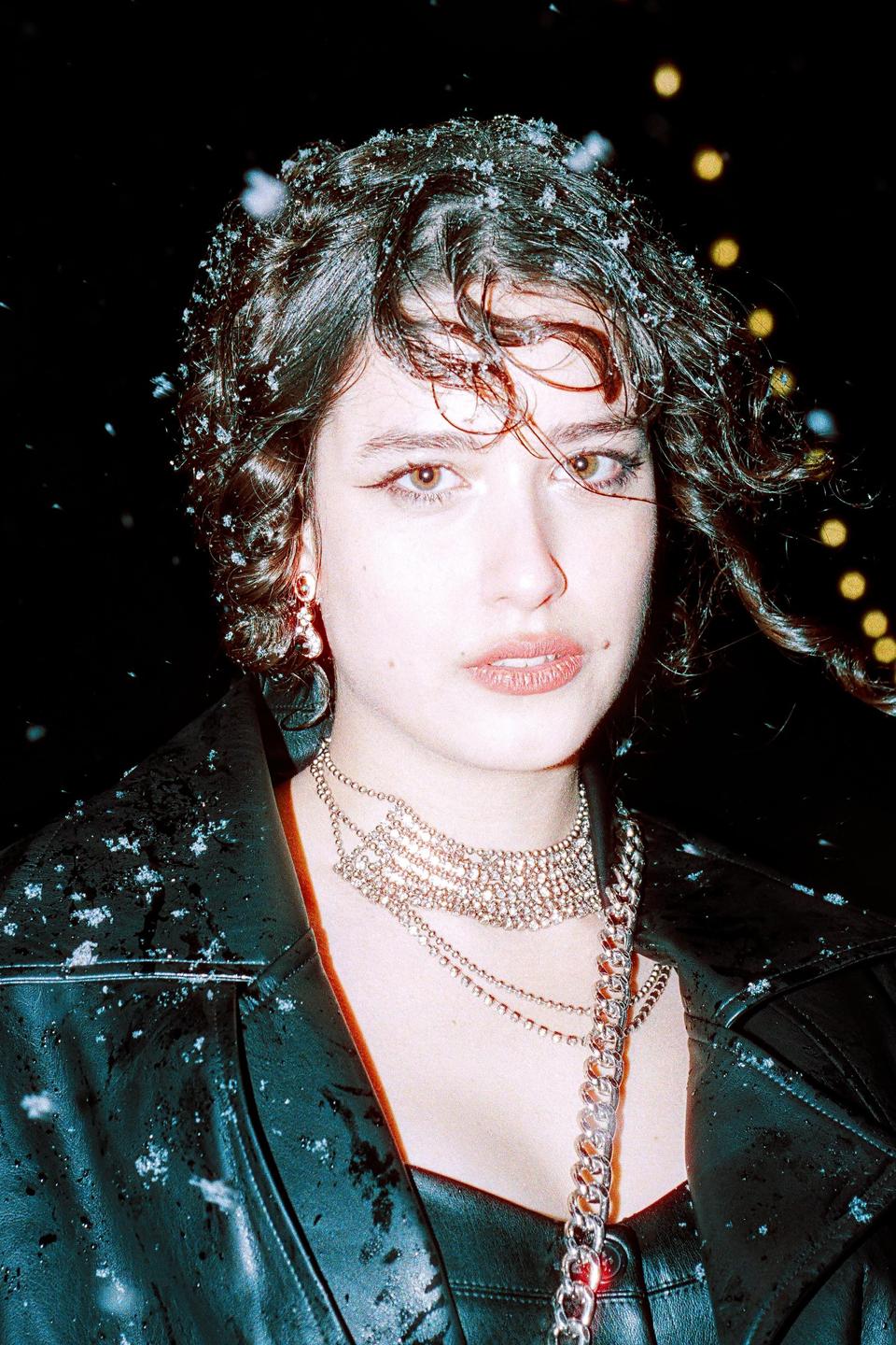 A woman with curly hair and a leather jacket is covered in snow at night.