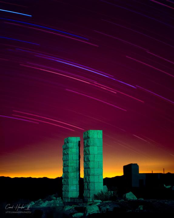 Two green stone pillars stand against a night sky with star trails and an orange glow on the horizon.