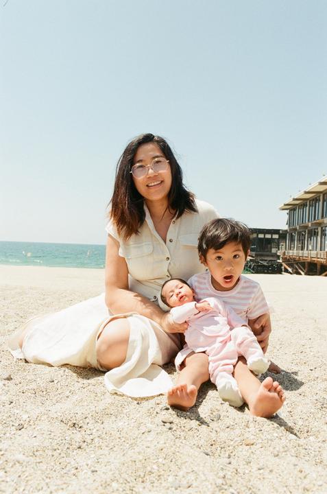 A woman sits on a sandy beach with a boy and a baby, with the ocean and a building in the background.