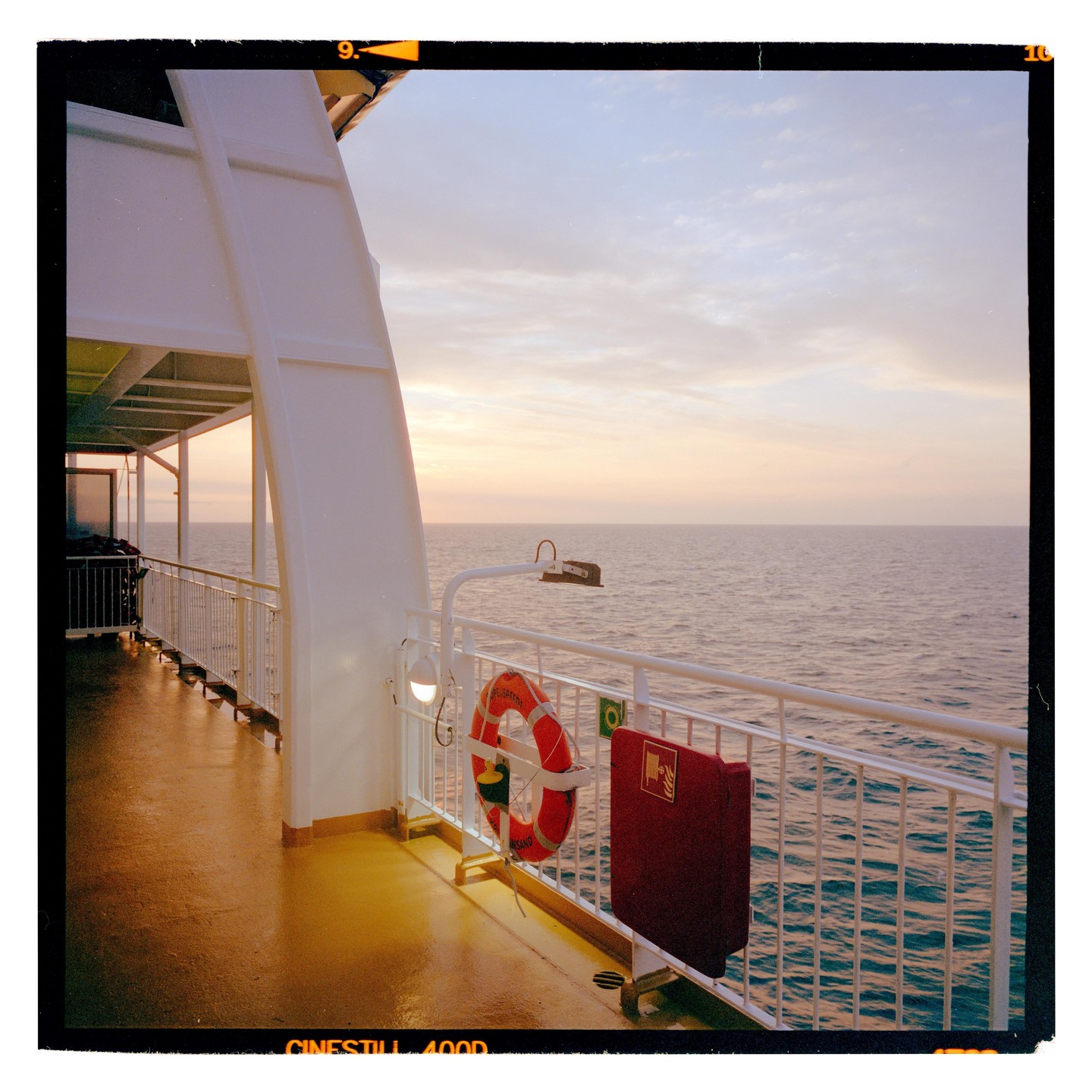 A ferry deck with a life preserver and fire hose box overlooks the ocean at sunset.