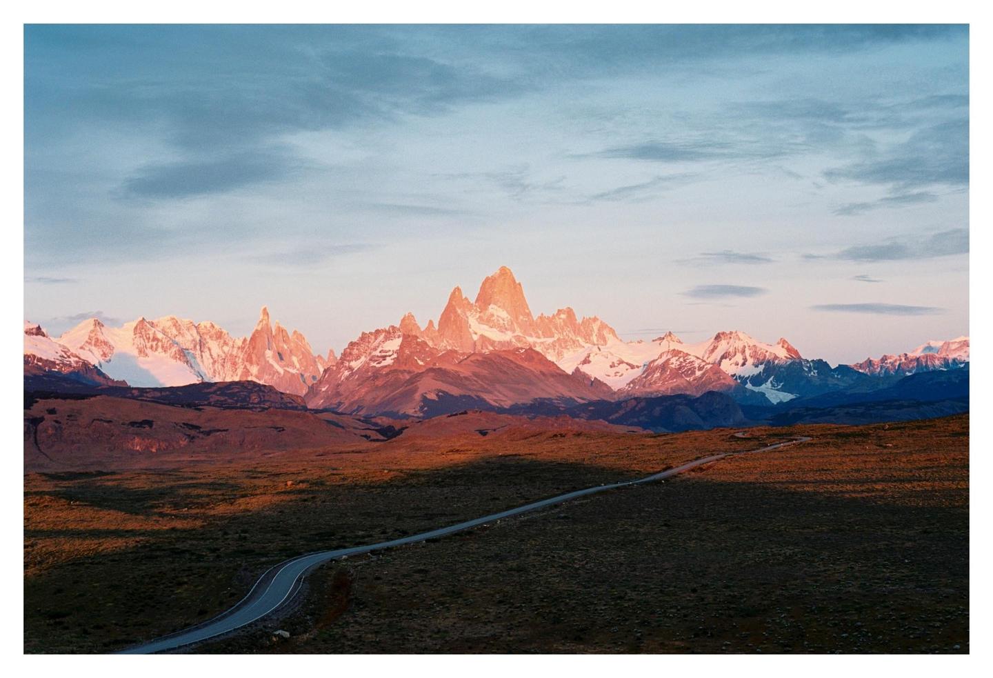 A winding road leads towards a mountain range with snow-capped peaks illuminated by the setting sun.
