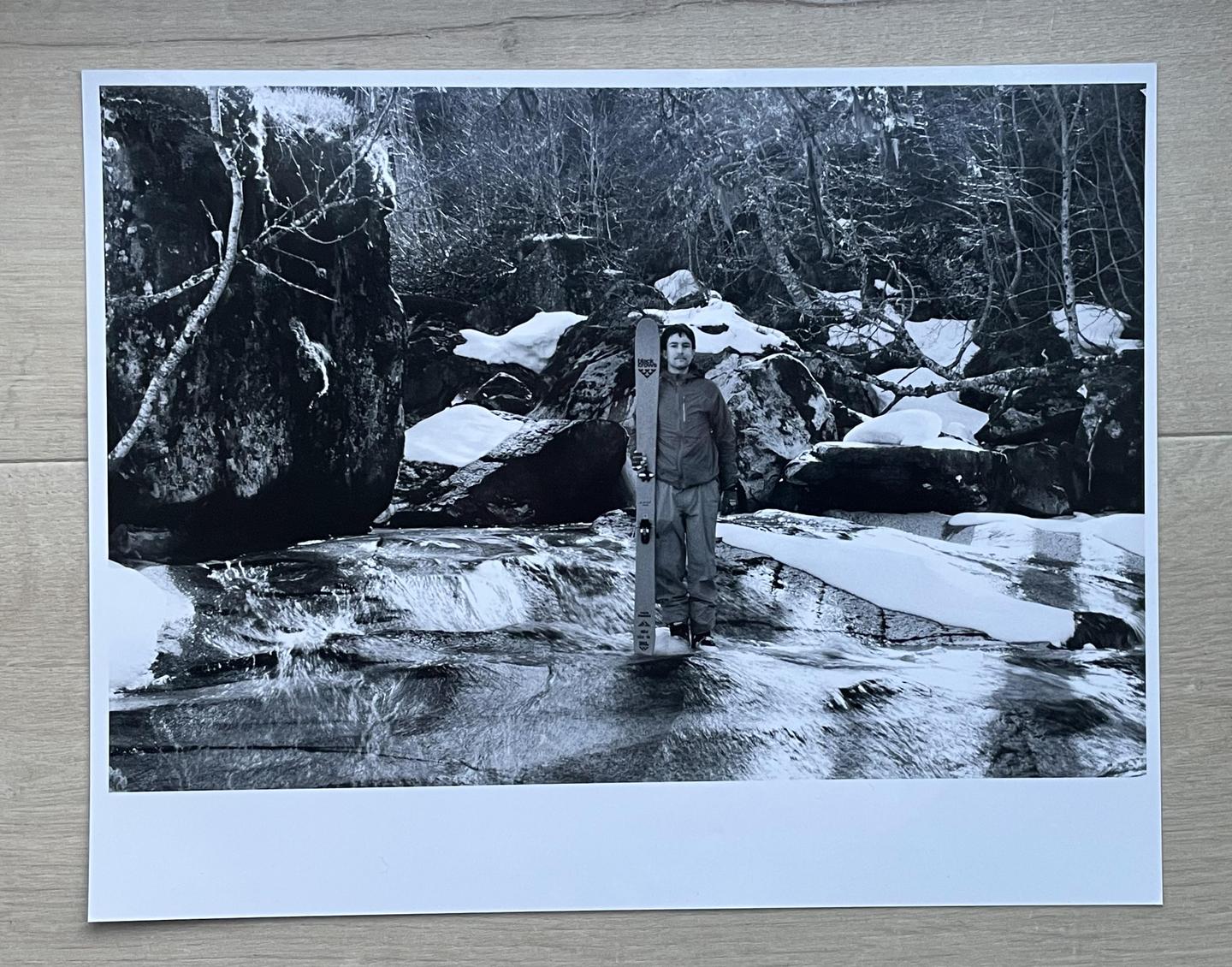 A man stands in a snowy river holding a ski, surrounded by rocks and trees.