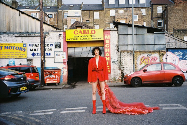 A person in a red jacket and red boots with a long red train stands in the middle of a street in front of a garage.