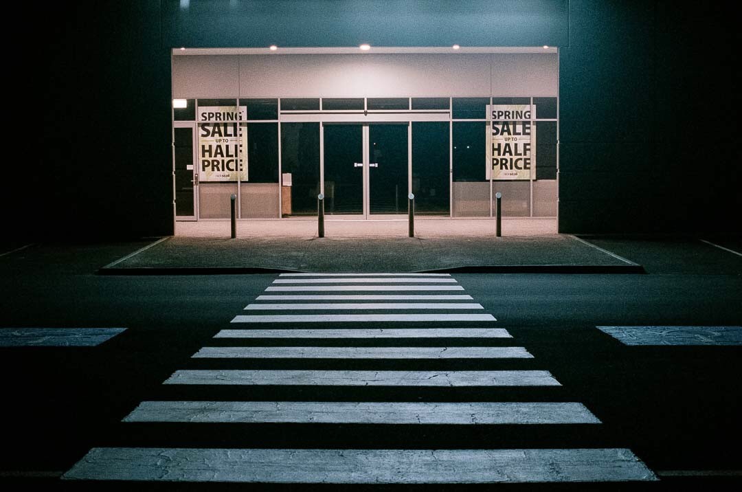 A crosswalk leads to the entrance of a store with "Spring Sale Half Price" signs in its windows at night.