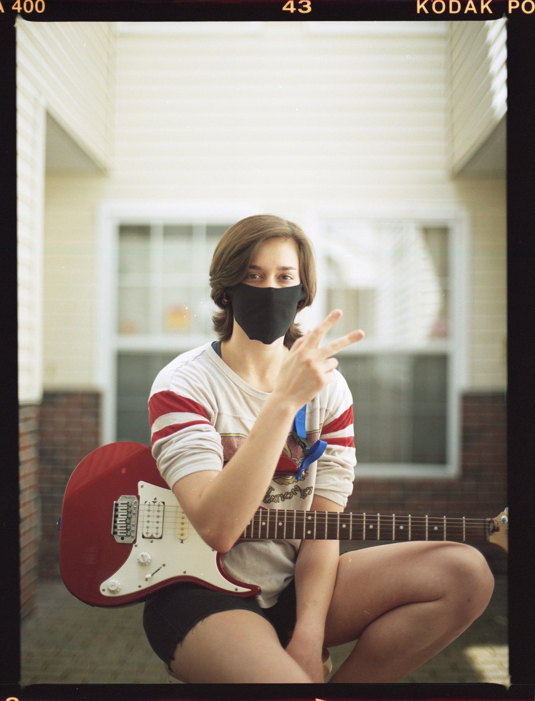 A woman wearing a face mask and holding a red electric guitar makes a peace sign.