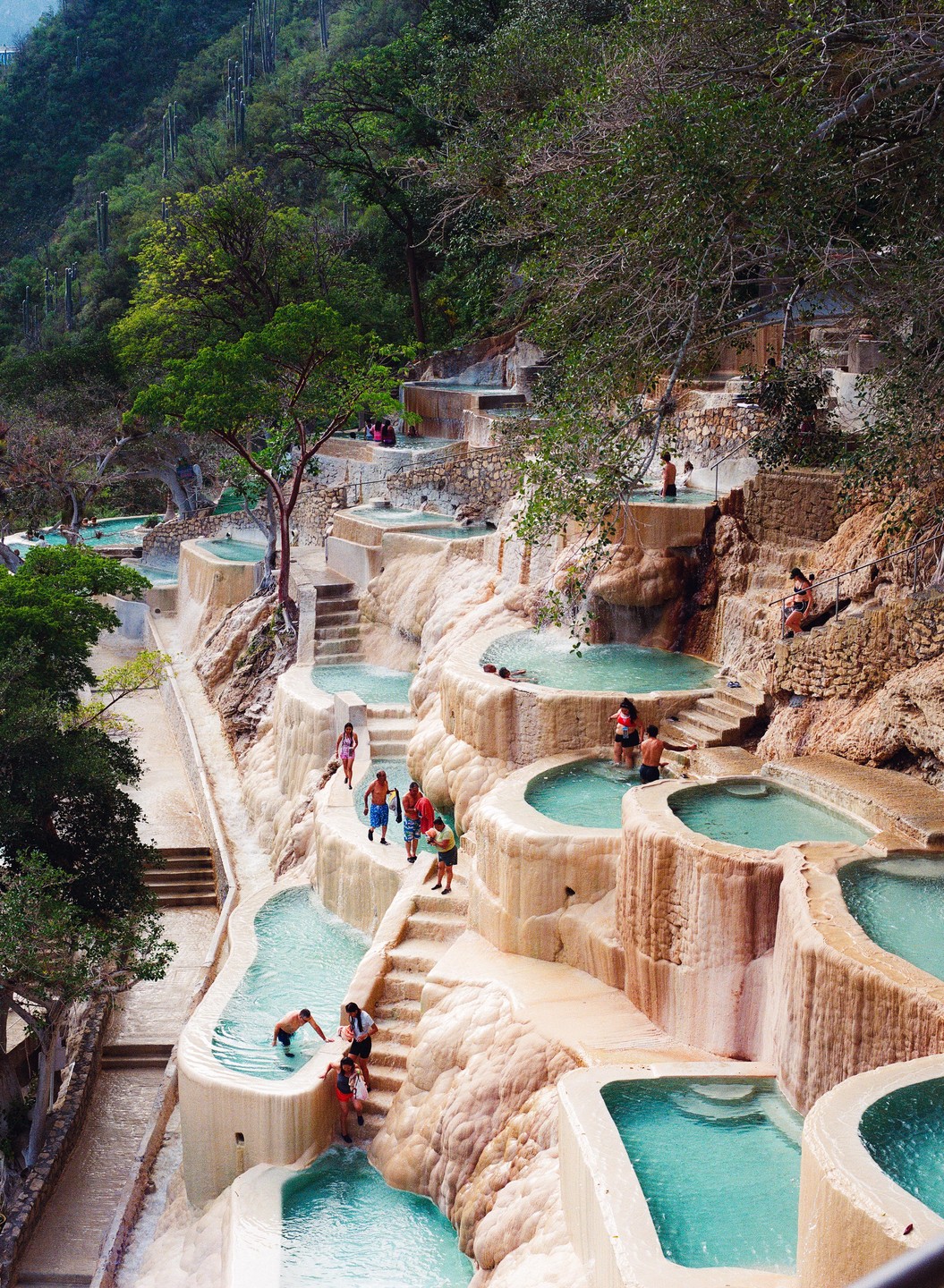 People are swimming and walking around terraced pools on a hillside.