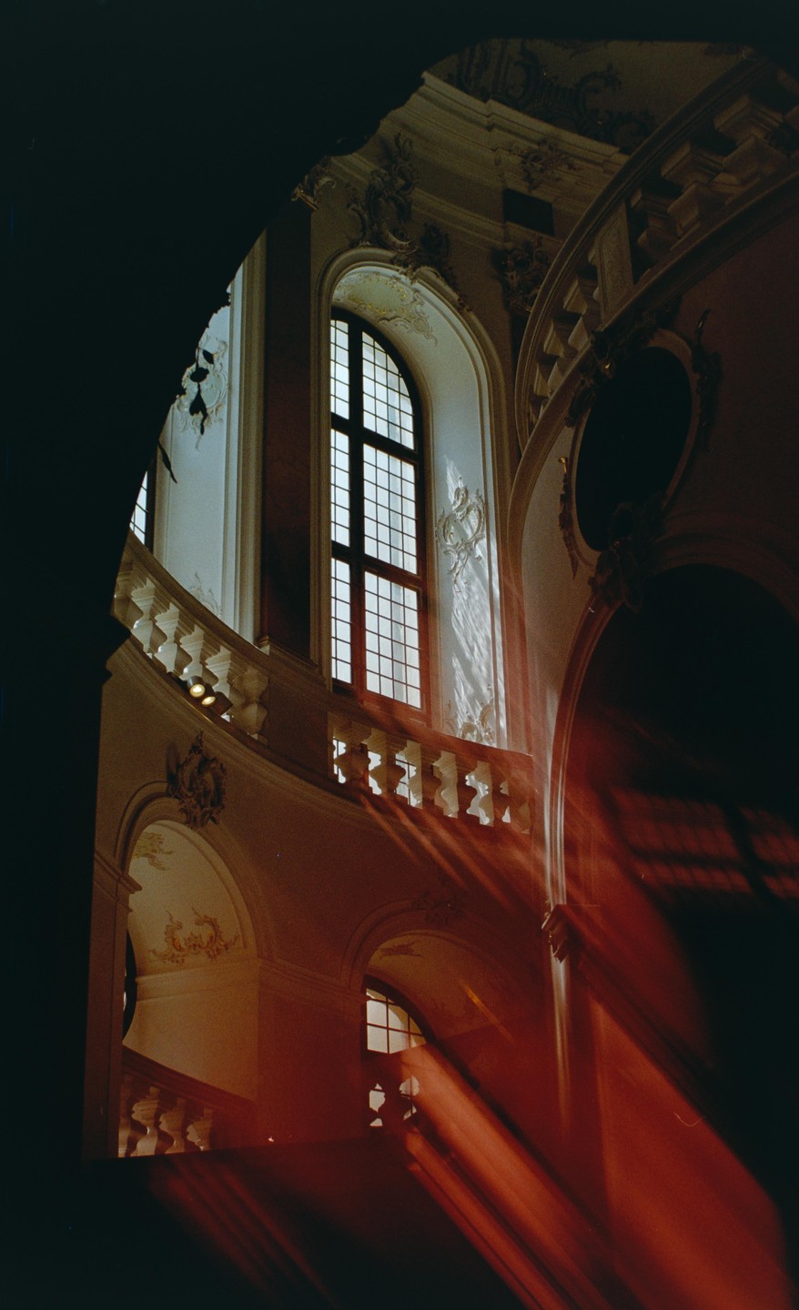 Sunlight streams through a tall arched window, illuminating a grand staircase with ornate balustrades and decorative walls.