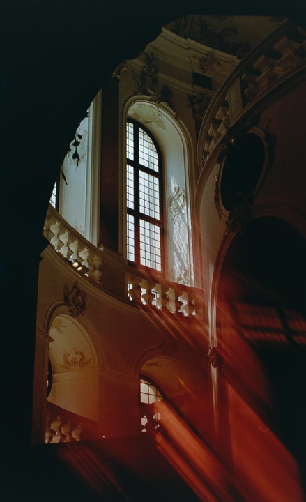 Sunlight streams through a tall arched window, illuminating a grand staircase with ornate balustrades and decorative walls.