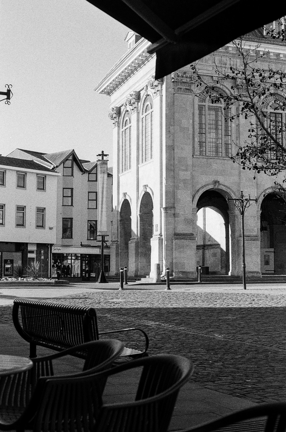 A black and white street view shows a large building with arches and columns, with a cafe terrace in the foreground.