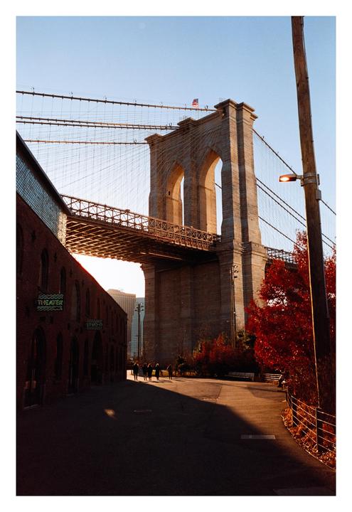 A street view of the Brooklyn Bridge with people walking on a path and a brick building on the left.