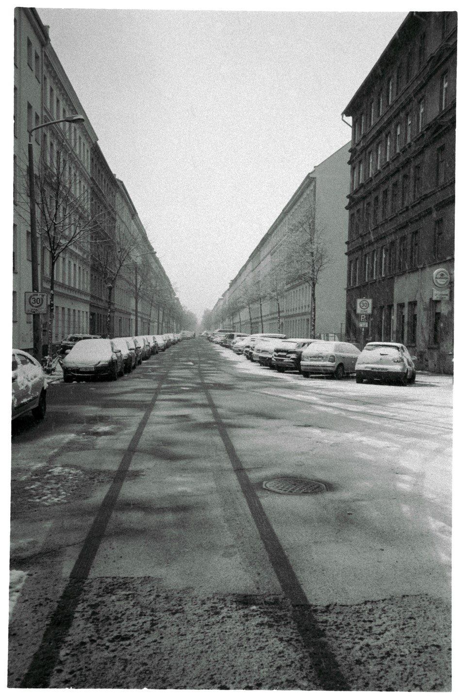 A snow-covered street with parked cars and tall buildings on either side stretches into the distance.