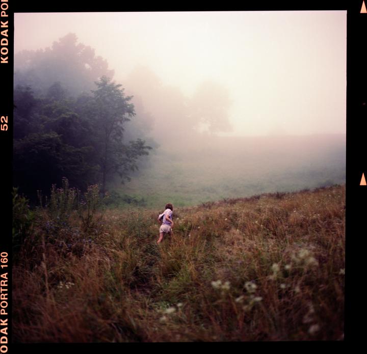 A child runs through a field of tall grass and wildflowers on a hazy day.