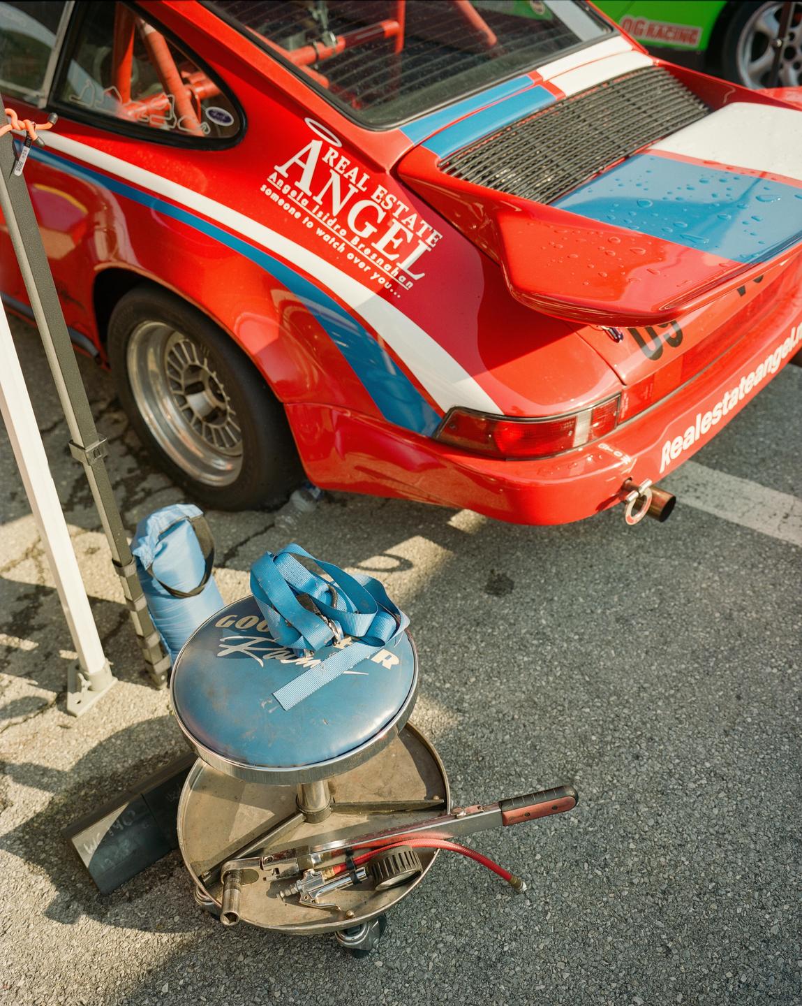 A red race car with blue and white stripes is parked on an asphalt surface next to a blue mechanic's stool with tools.