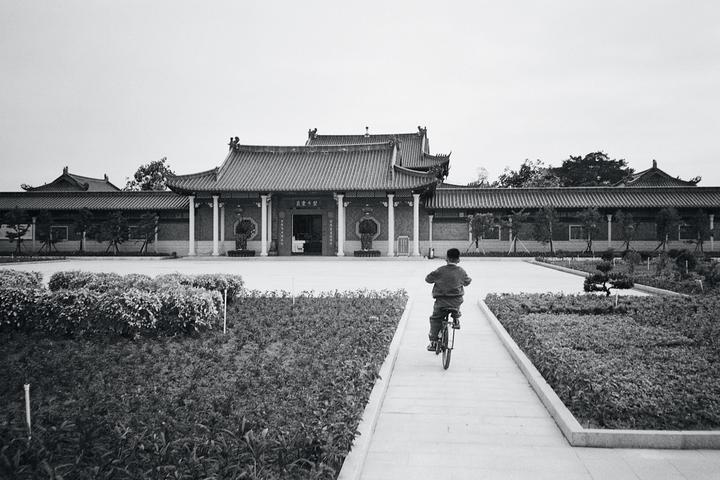 A boy rides a bicycle on a path leading to a traditional building with a tiled roof.