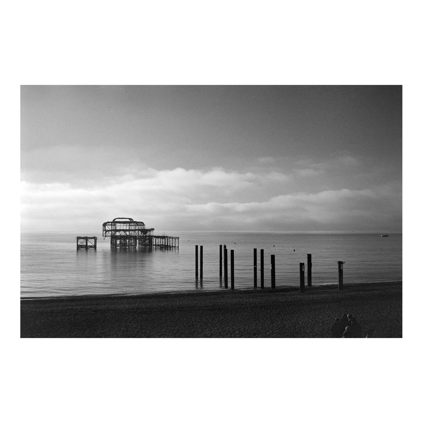 A ruined pier stands in the ocean with a pebble beach and several wooden posts in the foreground.