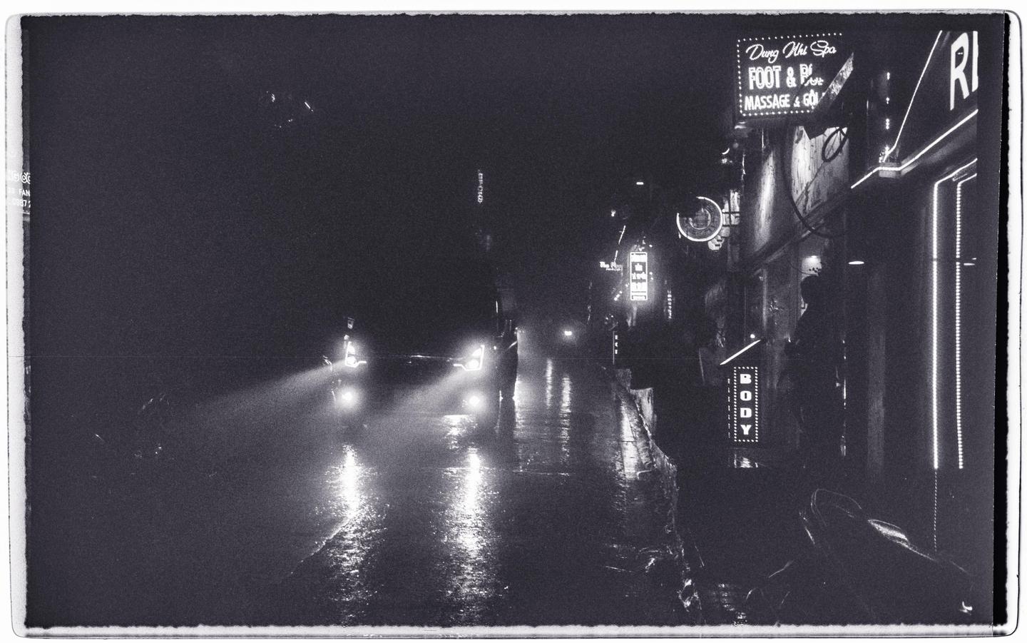 Headlights illuminate a wet street at night with neon signs on the right.