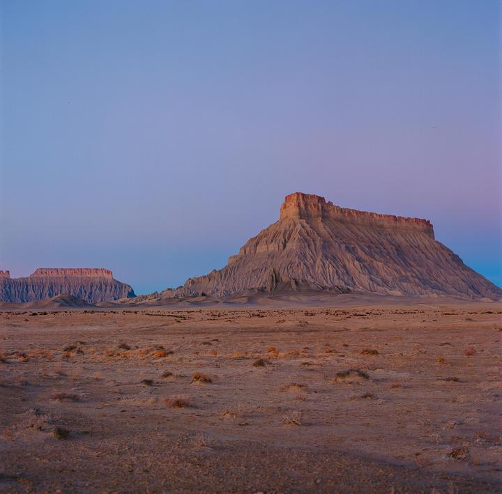 A vast desert landscape features a large mesa under a clear sky with a smaller mesa in the distance.
