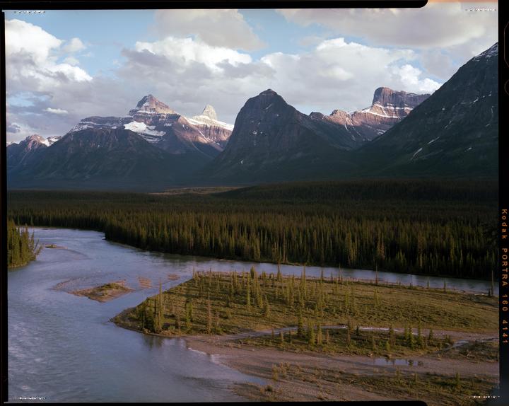 A wide river flows through a forest with snow-capped mountains in the background under a cloudy sky.