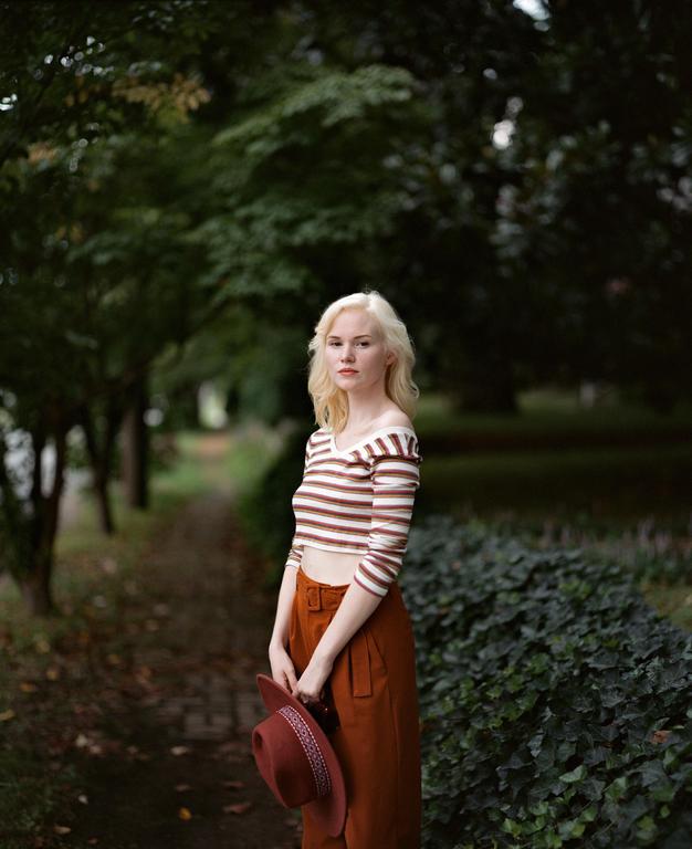 A woman with blonde hair stands on a path, holding a hat.
