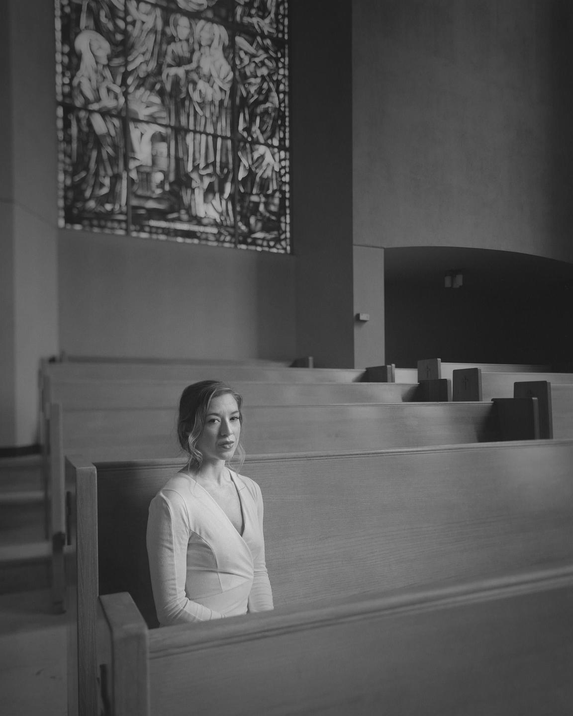 A woman sits in a pew in a church with a stained glass window in the background.