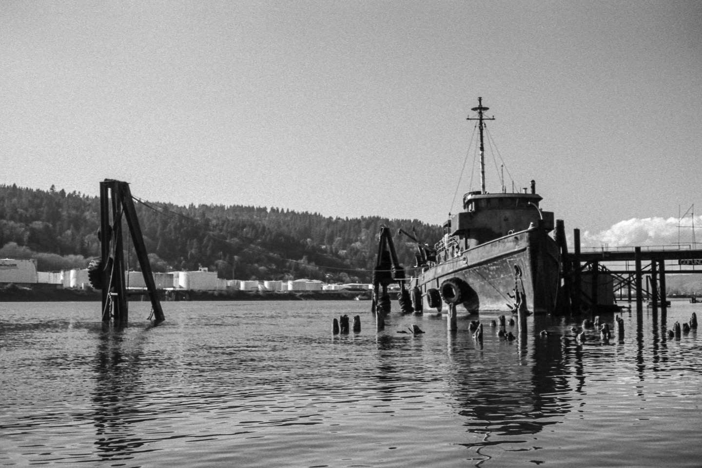 A large boat is docked next to a pier with a forested hill and industrial tanks in the background.