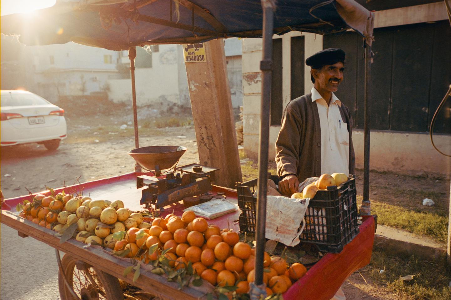 A man stands behind a fruit cart laden with oranges and guavas, with a white car visible in the background.