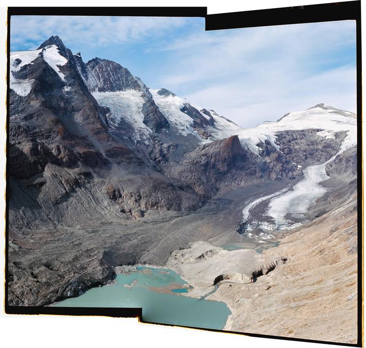 A panoramic landscape shows a glacier winding through a valley with snow-capped mountains and a turquoise lake.