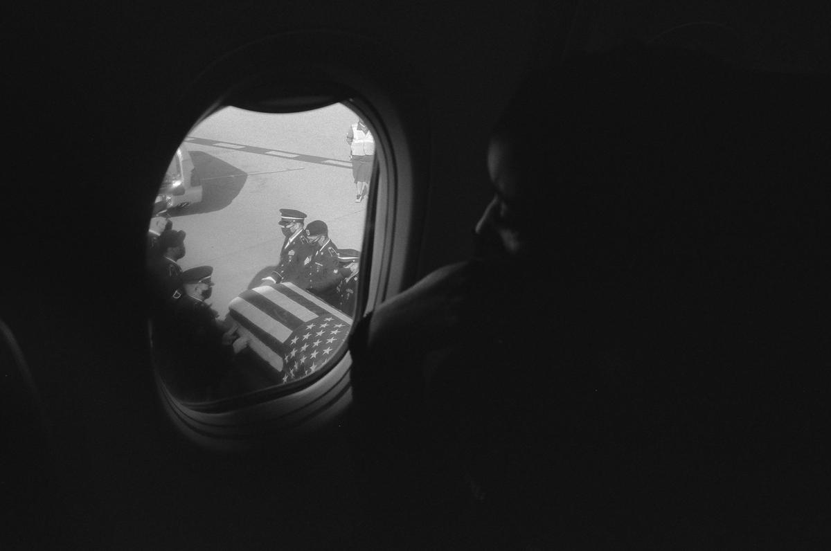 A person looks out an airplane window at a flag-draped coffin being carried by uniformed men on an airfield.
