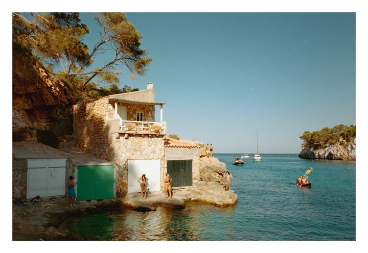 A group of people are swimming and relaxing by stone buildings on a rocky coast with boats in the water.