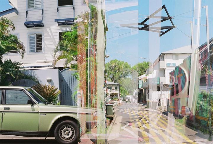 A double exposure shows a green car parked in front of a white building and a street with yellow markings.