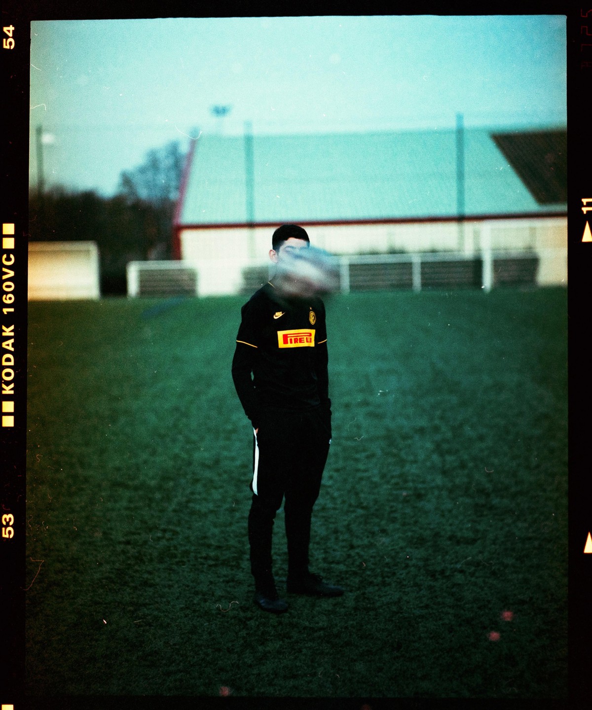 A man in a black soccer jersey stands on a green field with a blurred soccer ball in front of his face.