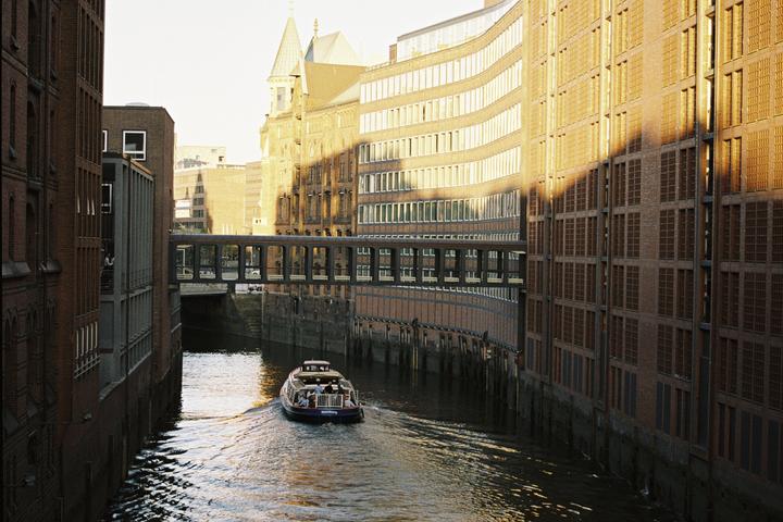 A boat travels down a canal between tall brick buildings with a pedestrian bridge overhead.