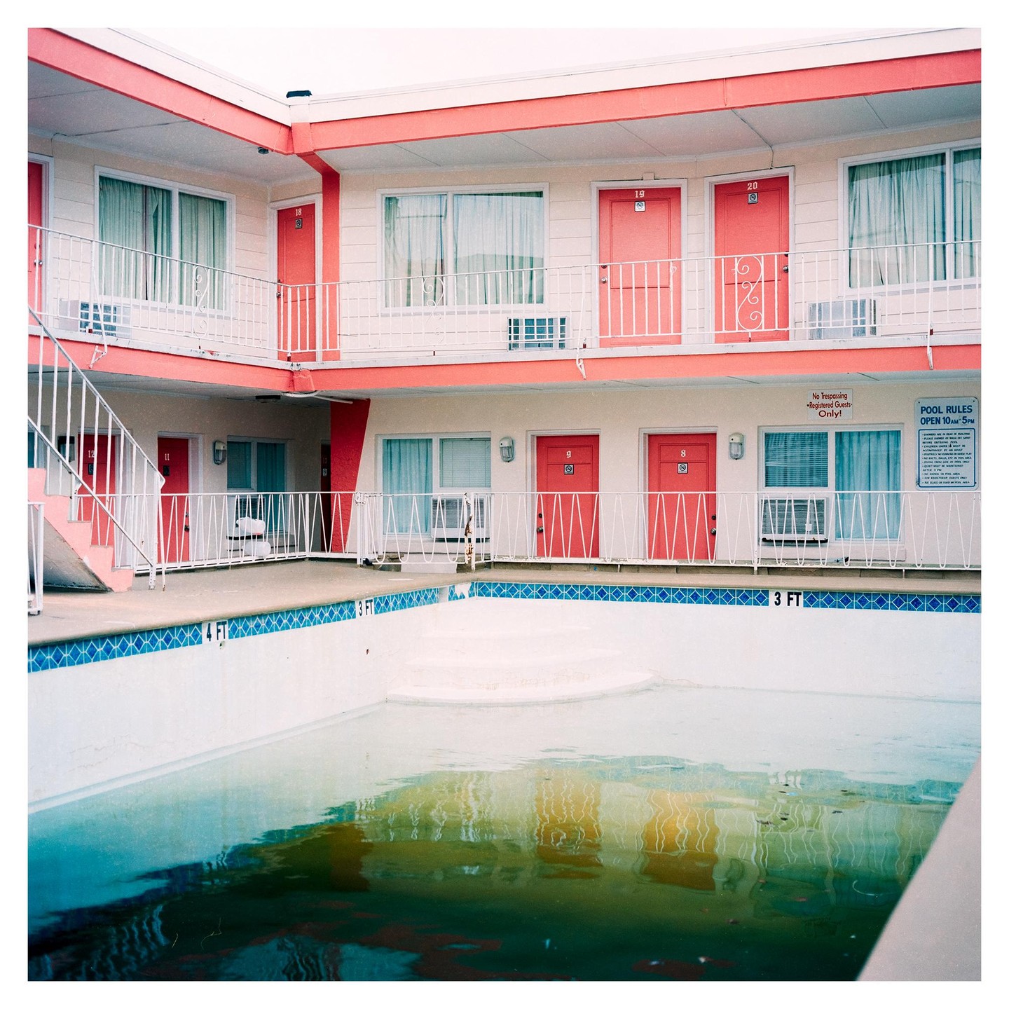 A motel with pink doors and railings surrounds a swimming pool with murky water.