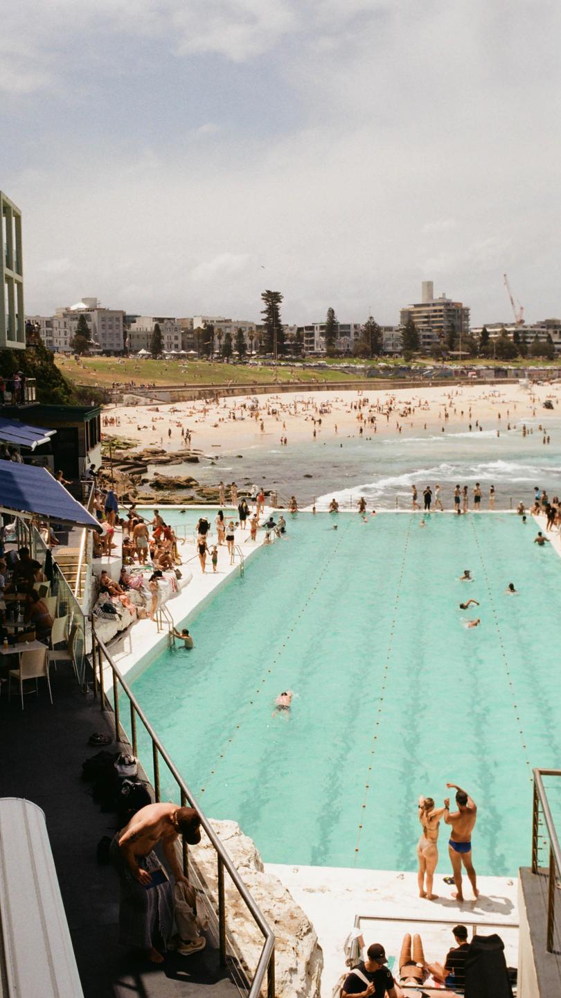 A large crowd of people are visible on a beach and in a swimming pool next to a town.