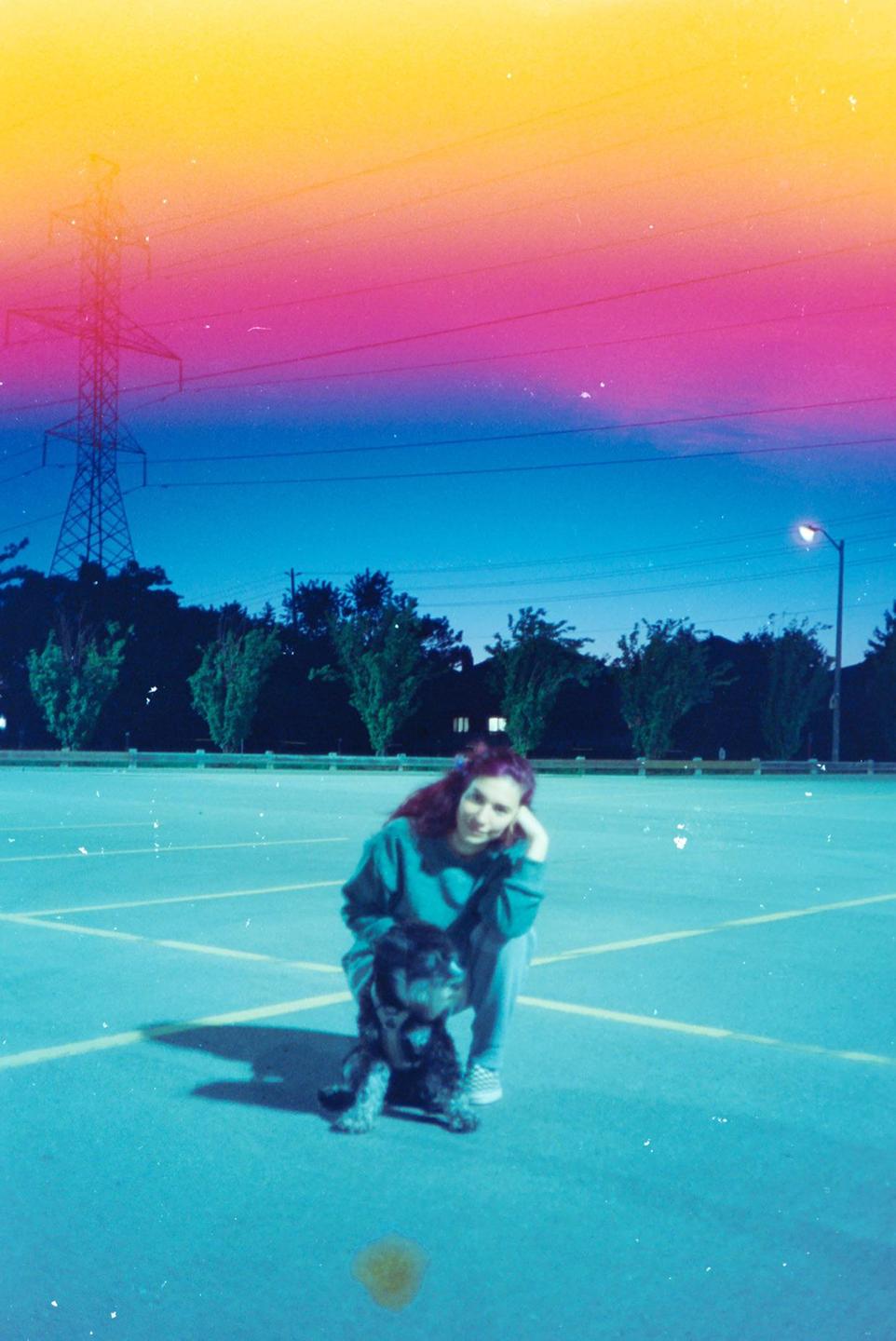 A woman and a dog are crouching on a parking lot at dusk, with a colorful light leak across the top of the image.