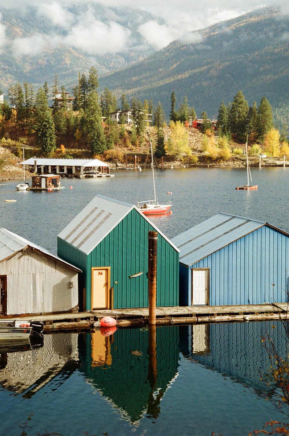 Three colorful boathouses stand on a dock with mountains and a lake in the background.