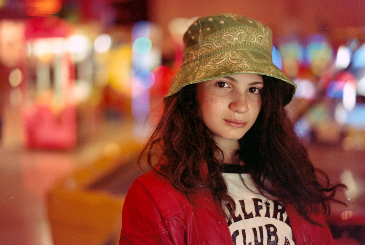 A girl with long hair and a bucket hat looks at the viewer, illuminated by colorful lights from an arcade game.